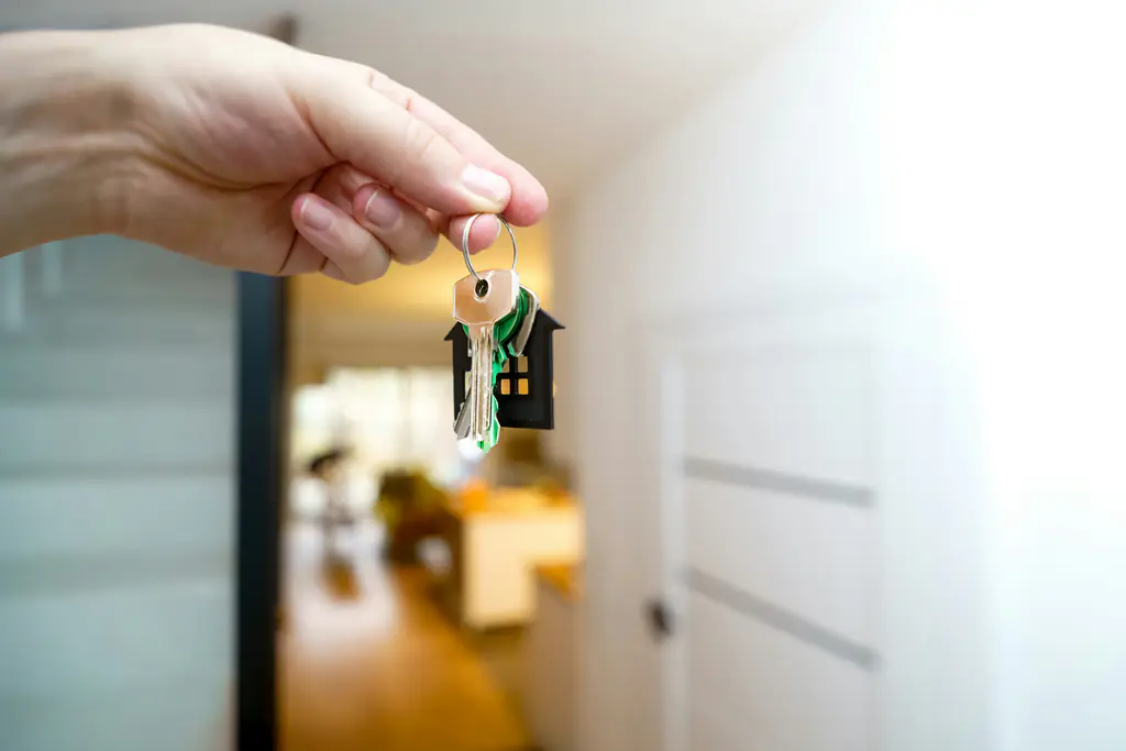 A hand holds a house-shaped keyring at an open front door, a lit hallway beyond